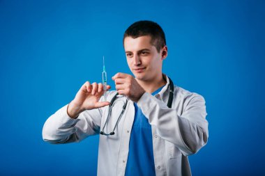 Steteskopla mavi arka planda izole edilmiş şırıngaya bakan odaklanmış doktor portresi