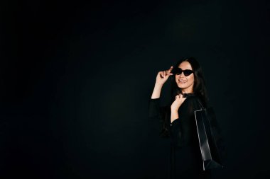 Güneş gözlüklü, siyah elbiseli ve sırtında alışveriş çantası olan güzel bir kız. Siyah üzerine izole edilmiş.