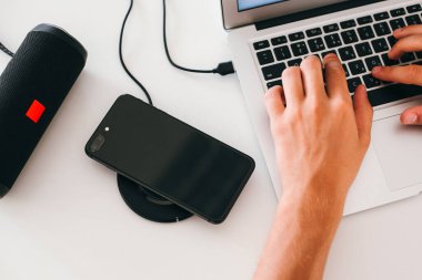 Adamın elleri dizüstü bilgisayarda, kablosuz hoparlörde ve kablosuz telefon şarjında çalışıyor.