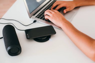 Adamın elleri dizüstü bilgisayarda, kablosuz hoparlörde ve kablosuz telefon şarjında çalışıyor.