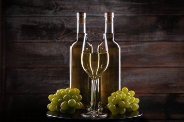 İki beyaz şarap şişesi, bir bardak ve ahşap bir arka planda üzüm.