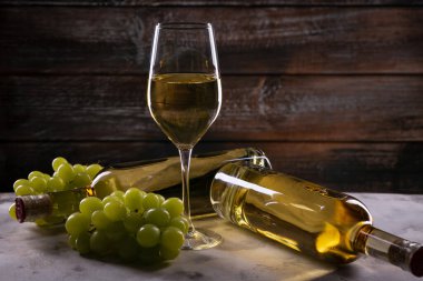 Üzümlü beyaz şarap kadehi ve iki şişe arkasında beyaz bir bez üzerinde ahşap bir zemin üzerinde yatıyor.