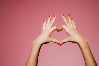 Parlak manikürlü bir kadının elleri pembe arka planda izole edilmiş aşk işareti parmakları