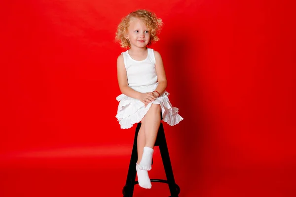 Modest little blonde girl in white dress sitting on chair isolated on red