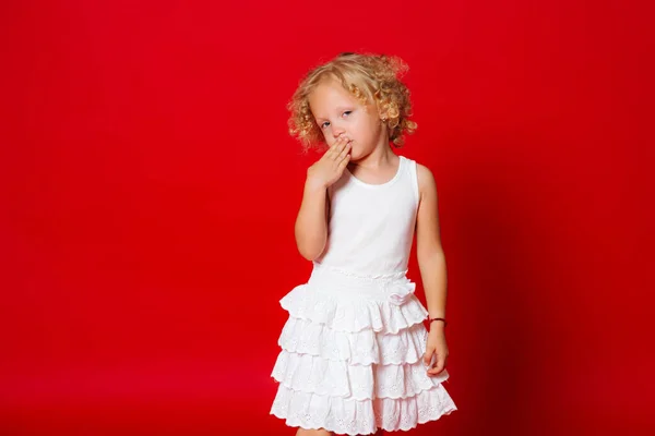 Pretty little blonde curly girl hides her face behind hand in white dress isolated on red