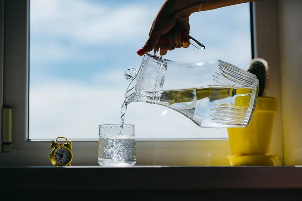 Woman\'s hands pouring water from a jug to glass on a windowsill