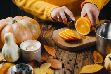 Manikürlü kadının elleri, sıcak şarap için tahta tahtada portakal keserken yakın plan görüntüsü. Balkabakları, yapraklar, masada mumlar. Sonbahar kipi.