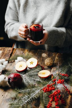 Gri kazaklı kadının ellerinde sıcak sıcak şarap. Masada duran güzel Noel süsleri: kürk ağacı dalları, kırmızı böğürtlen, elma, portakal, pamuk çiçeği. Şenlik havası.