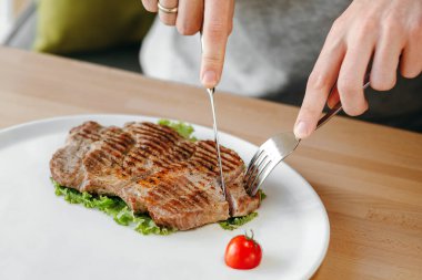 Adamın elleri bıçakla ızgara et kesiyor. Salata, vişneli domates ve biftek tabakta.