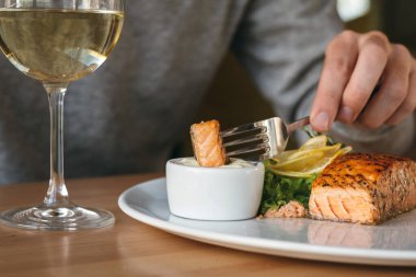 Somon balığı bifteğiyle çatal tutan bir adam. Adam somonu sosa batırıyor. Limon, salata ve biberiyeli ızgara somon bifteği.