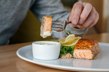 Somon balığı bifteğiyle çatal tutan bir adam. Adam somonu sosa batırıyor. Limon, salata ve biberiyeli ızgara somon bifteği.