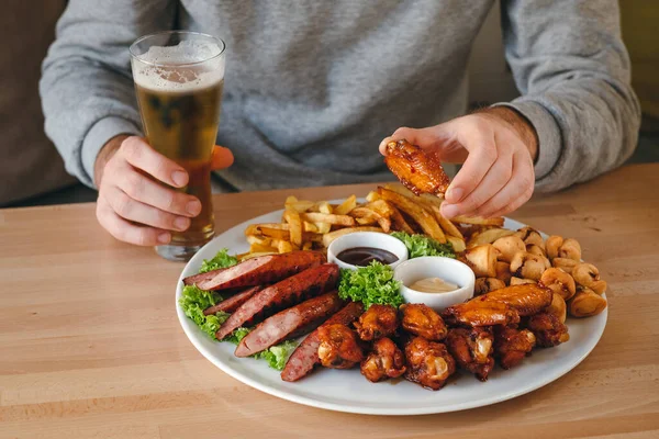 Man having dinner. Man\'s hands holding chicken leg and beer, large hot beer plate with sausages, fries, chicken and sauses on the table.