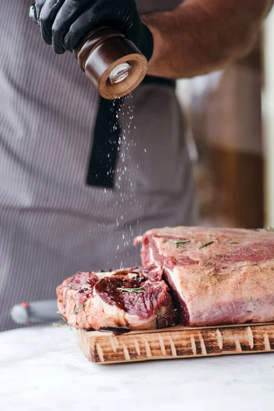 Chef adding salt to raw beef meat on chopping board, professional cook in gloves holding salt shaker, cooking steak.