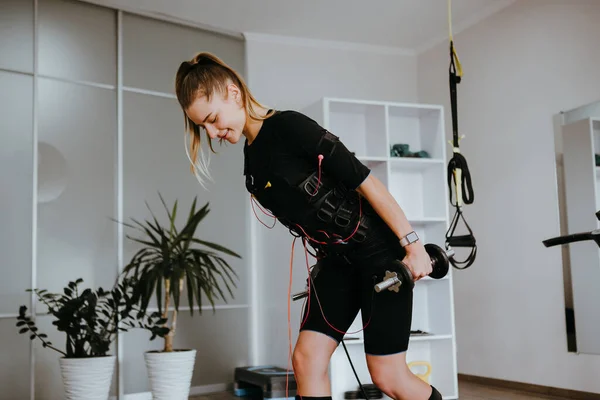 EMS training. Photo of young energetic woman lifting dumbbells in suit with cables.