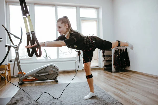 Woman doing exercises in EMS electro stimulation suit with cables, using TRX.
