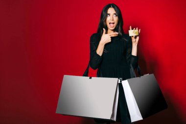 Koyu kırmızı arka planda altın bir kredi kartını işaret eden alışveriş torbaları taşıyan büyüleyici bir kadın..