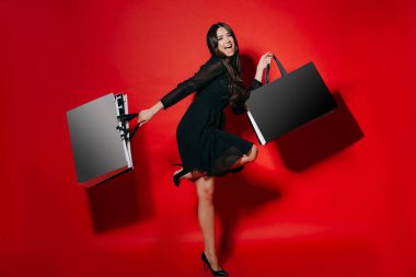 Siyah elbiseli bir kadın yüksek topuklu ayakkabıyla tek ayak üzerinde kırmızı arka planda alışveriş torbaları tutuyor. Alışveriş yaptıktan sonra alışveriş torbalarıyla dans eden heyecanlı kadın..