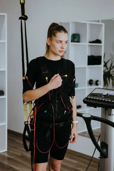 Woman standing next to EMS electro stimulation machine with cables, TRX hanging behind.