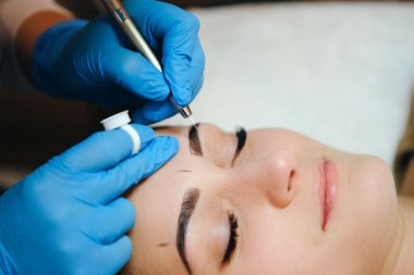 Güzellik salonundaki güzel esmer kadının kaşları için kalıcı makyaj. Eldivenli yakın güzellik uzmanı kaş yerine mikropla çalışıyor..