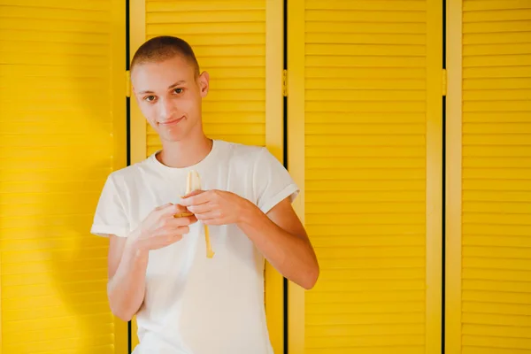 A hungry smiling guy in white t-shirt peeling a banana, on the yellow backgroung