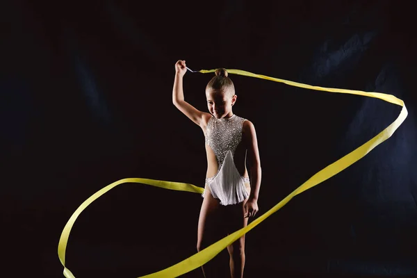 Girl gymnast in a white sparkling gymnastic swimsuit makes a snake in the air with a yellow ribbon on a black background