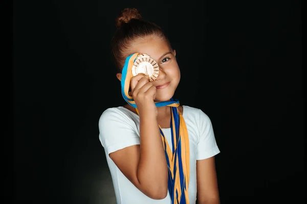 Portrait of little gymnast girl in white bodysuit with medals on her neck hiding happy face behind her medal isolated on black background