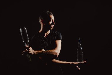 Yakışıklı barmenin dramatik portresi alkollü kokteylleri karıştırıyor. Profesyonel barmenlik hokkabazlığı ve şişesi ellerde ve siyahta izole.