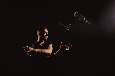 Yakışıklı barmenin dramatik portresi alkollü kokteylleri karıştırıyor. Profesyonel barmenlik hokkabazlığı ve şişesi ellerde ve siyahta izole.