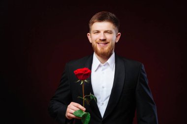 Takım elbiseli gülümseyen yakışıklı genç adam siyah bir arka planda kırmızı bir gül ile ayakta duruyor. Elinde gül tutan bir adamın romantik fotoğrafı..