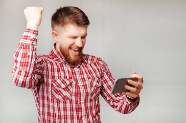 Gri arka planda heyecanlı sakallı bir adamın cep telefonuyla futbol maçı izleyip zaferi kutlaması..