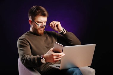 Kafası karışmış sakallı serbest çalışan sıradan giysiler içinde dizüstü bilgisayarına bakıyor ve elinde cep telefonu tutuyor. İş adamı uzaktan çalışıyor..