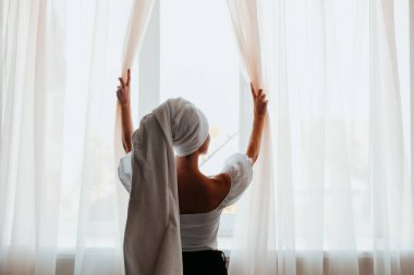 Kafasında havlu olan güzel bir kadın. Evde pencerenin yanında duruyor. Sabahları perdeleri açıyor..