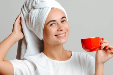 Gülümseyen genç bir kadın evde kahveyle eğleniyor. Beyaz tişörtlü ve kafasında kırmızı bardakla havluyla bir kadın fotoğrafı..