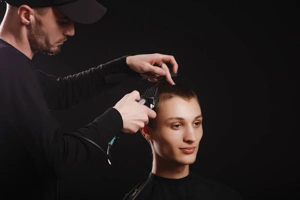 Close-up of barber in a black cap making haircut to attractive young man in barbershop on black background.
