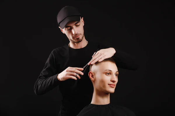 Barber in a black cap makes a haircut for a young man using scissors and comb, on black background.
