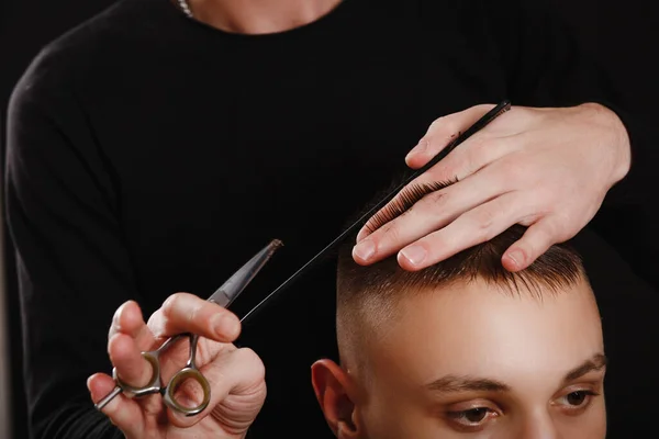 Close up of barber's hands using scissors and comb to make a haircut for a young man on black background.