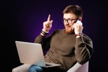 İş yerinde oturan, cep telefonuyla konuşan, parmakla işaret parmağını kaldıran, günlük kıyafetleri ve gözlükleriyle yakışıklı, genç, sakallı bir serbest çalışan..