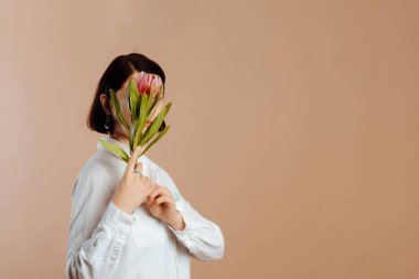 Geniş egzotik çiçekli, bej arka planda yüzünü saklayan genç bir kadının minimalist portresi..