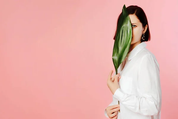 Portrait of young woman wearing white shirt, standing over pink background holding green palm leaf. Girl looking from behind green palm leaf.