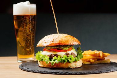 Domatesli sığır burger ve siyah tabakta patates kızartması ve arka planda yarım litre bira. Abur cubur. Fast food. Vücut pozitifi. Karantinadaki yiyecekler teslimattan. Menü için fotoğraf.