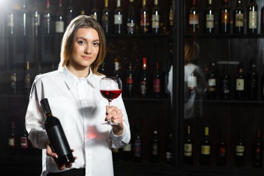 Sarışın, genç, güzel kız şarap garsonu elinde bardak, şişe ve bir bardak kırmızı şarap kokusu. Şarap aromasını takdir eder..