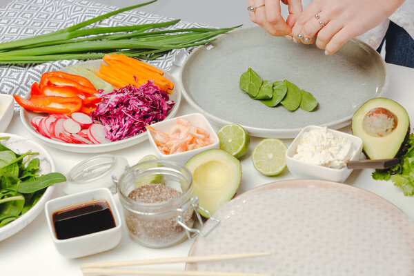 The process of cooking spring rolls - woman's hands putting basil on rice paper. Table with spring rolls ingredients and water with oranges.