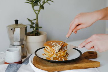 Kız mutfaktaki beyaz masada kahve makinesinin yanında bir parça krep kesiyor. Karantinada sabah kahvaltısı. Ev yapımı lezzetli krep tarifi..