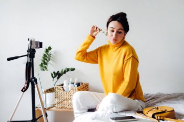 Genç esmer kız güzellik blogcusu beyaz arka plandaki odasında yüz masajı yapıyor. Blog çekmenin konsepti. Güzellik ve cilt bakımı.