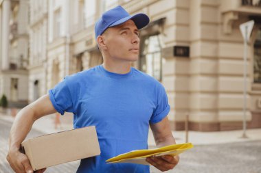Genç, neşeli teslimatçı. Elinde karton bir kutu var.