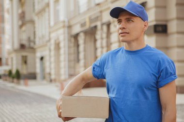 Genç, neşeli teslimatçı. Elinde karton bir kutu var.