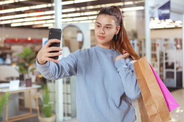 Güzel genç bir kadın alışveriş merkezinde alışveriş yaparken akıllı telefonuyla selfie çekiyor.