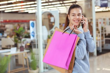 Hoş bir genç bayan telefonda konuşuyor, alışveriş merkezinde alışveriş torbalarıyla yürüyor.
