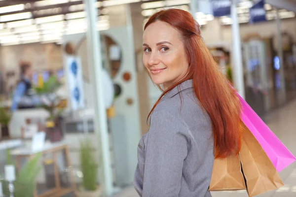 カメラに笑みを浮かべて豪華な赤い髪の女性は モールで買い物袋を運ぶ — ストック写真