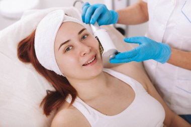 Happy young woman smiling to the camera while getting facial  massage by cosmetologist. Skin tightening, firming procedure concept